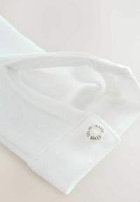 Valkoinen tekstiilikassi, jossa on teksturoitu kangaspinta, kaarevat kahvat ja pyöreä hopeinen logo sivussa.