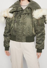 Chaqueta bomber verde oliva con un cuello texturizado y ribete de pelo, y una cinturilla ajustada. Presenta grandes botones y bolsillos con cremallera.