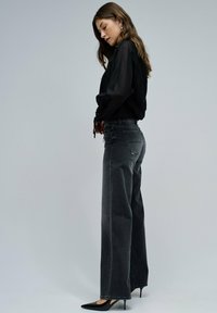 Mujer de pie de lado, vistiendo una blusa negra de manga larga, jeans oscuros de talle alto, tacones negros de punta y pendientes de plata.