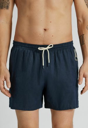 Mann trägt marineblaue Shorts mit Kordelzug und einer kleinen Metallklammer an der Seite, steht vor einem einfachen Hintergrund.
