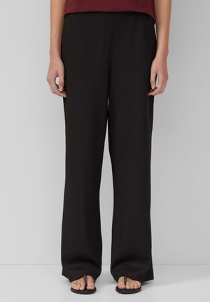Pantalons noirs à jambe large en tissu texturé, affichant une coupe décontractée et des détails de plis subtils. Associés à des sandales plates.