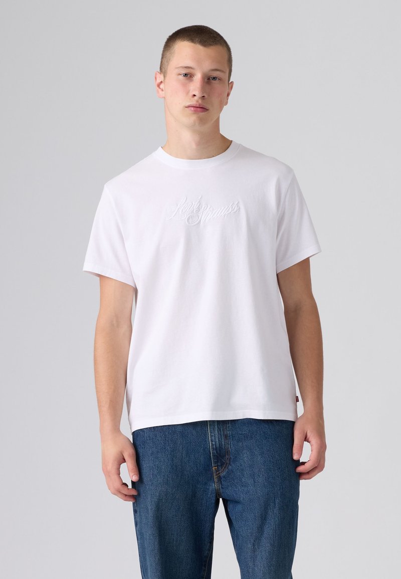 Young man wearing a plain white short-sleeve t-shirt with subtle script and blue jeans, standing against a plain light gray background.