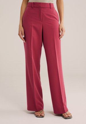 Kobieta w różowych spodniach z wysokim stanem i szerokimi nogawkami, z przodu plisowanych, oraz w sandałach na otwartym palcu z nadrukiem węża na jasnym tle.