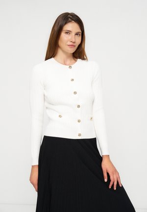Frau trägt eine weiße Strickjacke mit goldenen Knöpfen und einen langen schwarzen Rock, steht vor einem einfarbigen hellen Hintergrund.