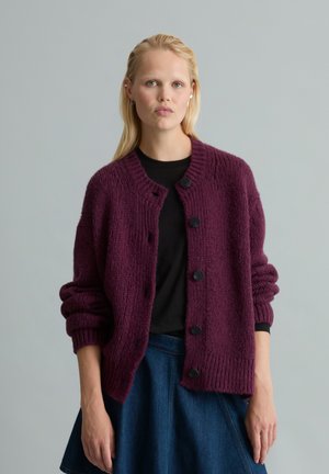 Frau mit blondem Haar trägt eine lila grob gestrickte Strickjacke über einem schwarzen Shirt und einem blauen Rock, stehend vor einem einfarbigen grauen Hintergrund.