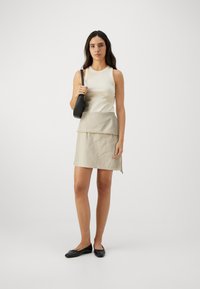 Beige ermeløs topp parret med en lys beige skjørt med bølgekant, og svarte flate sko. Modellen bærer en svart veske.