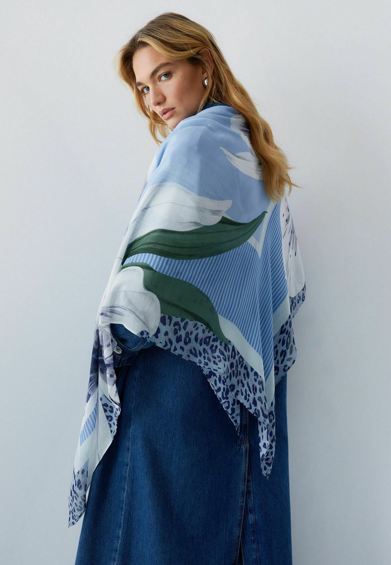 Femme aux longs cheveux blonds portant un manteau en denim bleu et une grande écharpe fleurie bleu clair drapée sur ses épaules, regardant par-dessus son épaule.