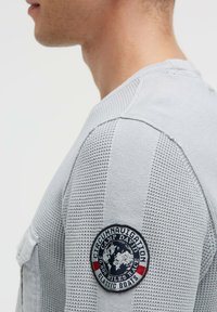 Graues Langarmshirt mit strukturiertem Strickmuster und einem gestickten Aufnäher mit einer Weltkugel und Schriftzug in kontrastierenden Farben auf der Schulter.