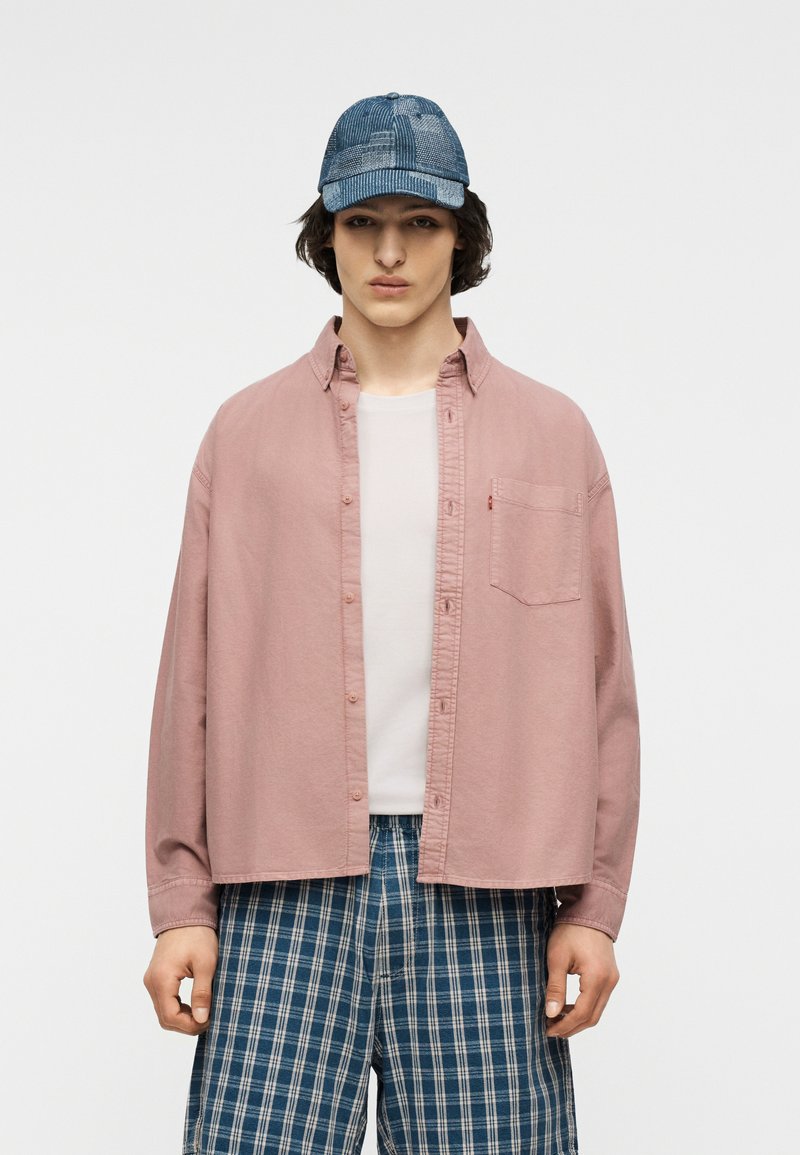 Junger Mann mit einer blauen, gemusterten Kappe, locker sitzendem rosa Hemd über einem weißen T-Shirt und blauen karierten Shorts, der vor einem schlichten Hintergrund steht.