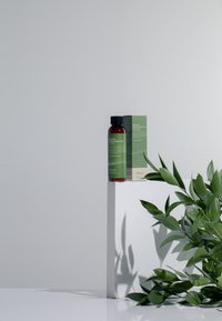 Brun flaska med svart kork bredvid grön låda, båda märkta "Benton Hyaluron Grönt Te Lotion," på en vit pelare med gröna blad.