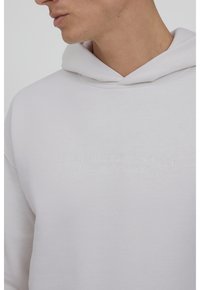 Sweat à capuche gris clair en coton doux, avec une grande poche avant et un logo de marque embossé. Le design comprend une capuche avec cordon de serrage.