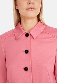 Veste rose avec un col, des boutons noirs, une texture lisse et des poches latérales ; conçue avec une coupe ajustée et des détails minimaux.
