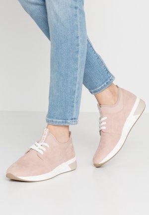 Personne portant un jean bleu clair et des baskets en suède rose clair avec des lacets blancs et des semelles en caoutchouc beige, debout sur un sol gris.