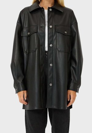 Personne portant une chemise-veste en cuir noir ample à boutons avec deux poches poitrine et manches longues par-dessus un haut blanc et un pantalon noir.