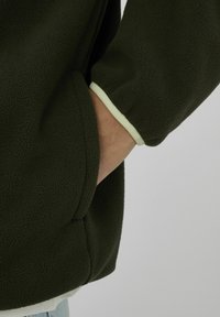 Veste polaire verte à surface texturée, avec une bordure vert clair le long des poignets et une main glissée dans la poche.