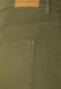 Tissu en denim vert olive avec une texture lisse, présentant une étiquette en cuir marquée "THE DENIM STORY" et des détails de coutures structurées.