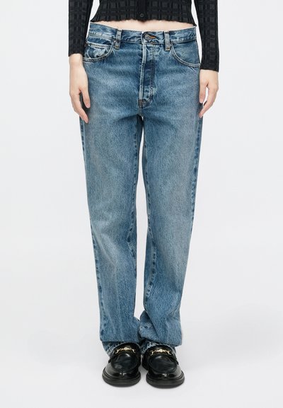 Jean en denim bleu clair avec une coupe décontractée, comportant cinq poches, une fermeture à boutons et des détails de surpiqûre visibles. Associé à des mocassins noirs.