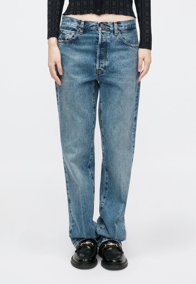 Jean en denim bleu clair avec une coupe décontractée, comportant cinq poches, une fermeture à boutons et des détails de surpiqûre visibles. Associé à des mocassins noirs.