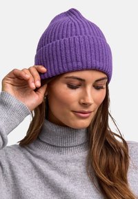 Woman wearing a purple knitted beanie and a grey turtleneck sweater, touching the edge of her hat with her right hand.