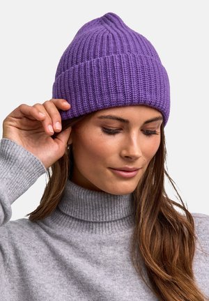Donna che indossa un berretto a maglia viola e un maglione grigio a collo alto, mentre tocca il bordo del suo cappello con la mano destra.