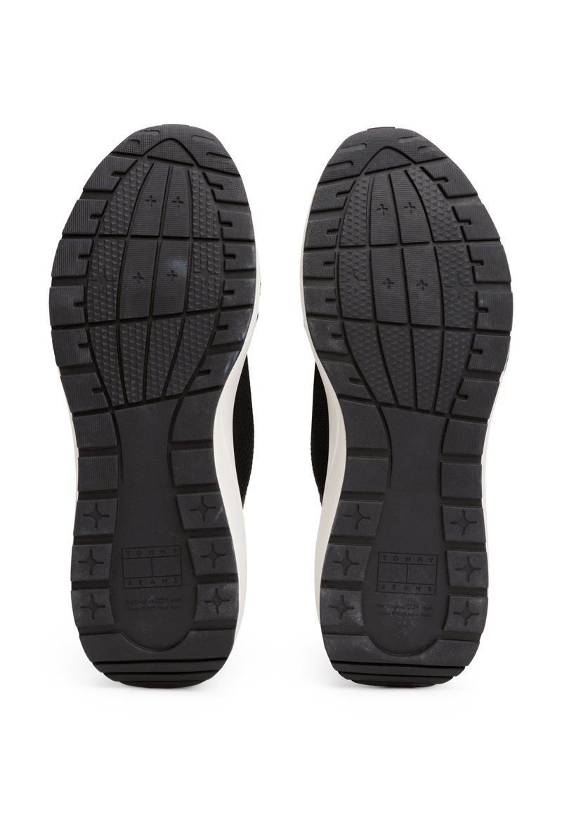 Semelles de chaussures en caoutchouc noir avec des motifs de relief profonds et une semelle intermédiaire blanche. Présente un logo rectangulaire. Surface texturée pour une meilleure adhérence.