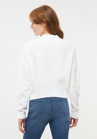 Biały sweter dzianinowy o teksturowanym wzorze, z ribbowanym dołem i akcentami w splocie warkoczowym na rękawach, noszony z niebieskimi dżinsami.