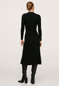 Femme avec les cheveux en chignon portant une robe midi noire à manches longues et des bottes noires à talons hauts, se tenant de dos sur un fond uni.