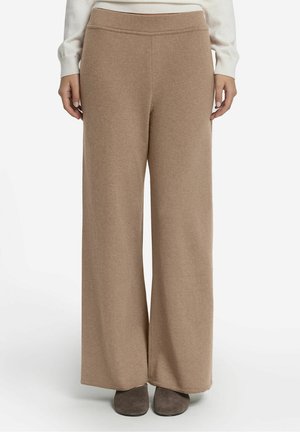Vrouw draagt beige, hoge taille, wijde gebreide broek gecombineerd met bruine schoenen en een wit shirt met lange mouwen.