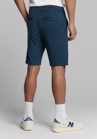 Navyblauwe shorts met een gladde textuur, voorzien van twee achterzakken en een elastische tailleband, gedragen met witte sokken en sneakers.