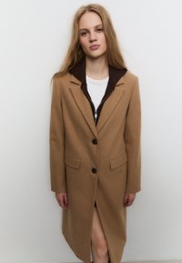 Cappotto in misto cammello con colletto marrone con zip, doppi bottoni e tasche laterali; lunghezza al ginocchio con uno spacco frontale per facilitare il movimento.