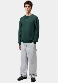Sudadera de manga larga color verde oscuro con puños acanalados, combinada con pantalones de mezclilla oversize en gris claro. Zapatos negros con superficie texturizada.