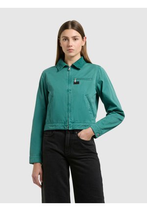 Kobieta ubrana w zielononiebieską kurtkę na zamek z kołnierzem i czarne spodnie, stojąca na jasnoszarym tle, z ręką w kieszeni.