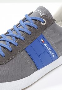 Baskets grises avec lacets bleus, arborant une bande en cuir bleue et le logo "HILFIGER". La chaussure possède une tige en tissu texturé et une semelle blanche.