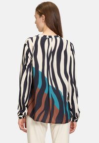 Blouse à manches longues avec un fond crème, ornée de motifs en vagues audacieux de couleur marine, sarcelle et marron. Tissu doux, coupe décontractée et poignets boutonnés.