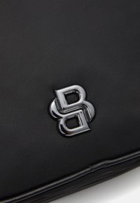 Matériau noir texturé avec un logo argenté brillant représentant des lettres entrelacées "B" sur la surface, mettant en valeur un design épuré et une finition haut de gamme.