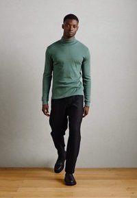 Grünes langärmliges Turtleneck-Oberteil, figurbetontes Design, kombiniert mit schwarzen Hosen und schwarzen Schuhen, vor einem neutralen Hintergrund mit Holzfußboden.