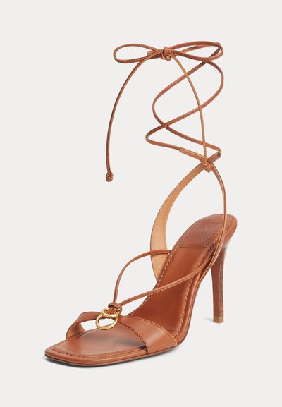 Καφέ δερμάτινο σανδάλι με ψηλό τακούνι, λεπτά λουράκια που τυλίγονται γύρω από τον αστράγαλο και μικρές χρυσές κρίκους ως διακοσμητικό στο μπροστινό λουράκι.