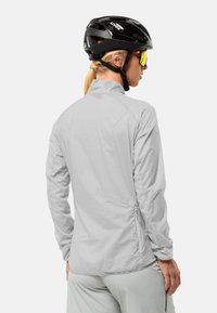 Lichtgrijze fietsjacket met lange mouwen, hoge kraag en ritsen aan de zijkanten. Draagt een zwarte helm en spiegelachtige zonnebril.