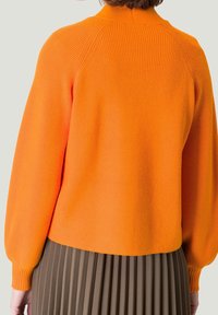 Jasnopomarańczowy sweter z dzianiny o prążkowanej fakturze, okrągłym kołnierzykiem i bufiastymi rękawami, zestawiony z plisowaną brązową spódnicą.