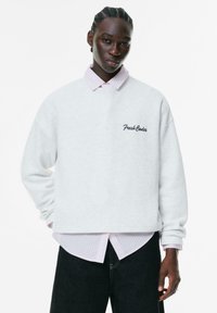 Grå crewneck sweatshirt med "Fresh Codes" broderi, lagd over en lyserød stribet skjorte, parret med mørke jeans. Glat tekstur.