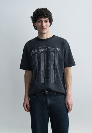 Ung mand med krøllet hår iført en mørk oversized t-shirt med "Infinite Secret Tour '99" og turnédatoer, kombineret med mørke jeans, stående foran en ensfarvet baggrund.