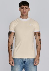 Beige aansluitend T-shirt met een witte geribbelde kraag en mouwaccenten. Heeft een klein logo op de borst. Gegd aan met lichtblauwe versleten jeans.
