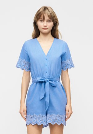 Nuori nainen pukeutuneena vaaleansiniseen, nappikiinnitteiseenhaalariin, jossa on vyötärönauha sekä liepeiden ja shortsejen helmassa aaltoilevaa kirjailua.