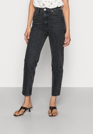 Kvinde iført sort højtaljede jeans, hvid blomstret top og sorte hælsandaler står på et gråt gulv mod en ensfarvet baggrund.