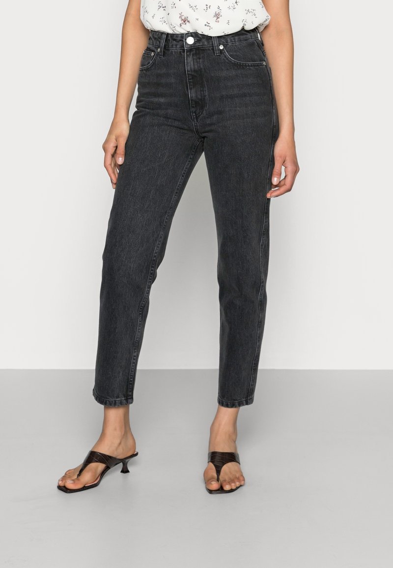 Femme portant un jean noir taille haute, un haut blanc à fleurs et des sandales à talons noires, debout sur un sol gris devant un fond uni.