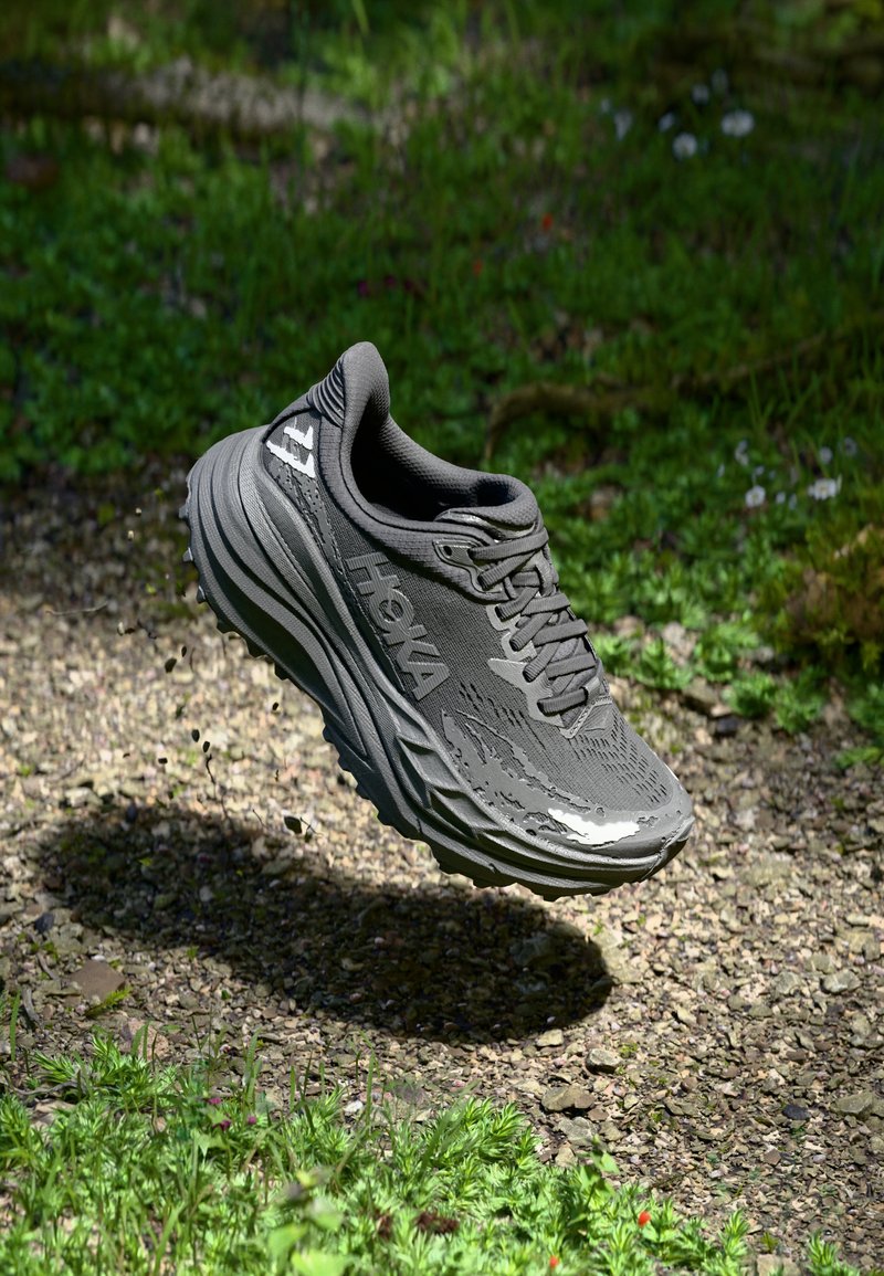 Scarpa atletica HOKA grigia con tomaia testurizzata, lacci visibili e una suola spessa e ammortizzata, sospesa sopra una superficie di ghiaia e erba.