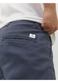 Pantalon en denim bleu marine avec une poche arrière ornée d'un accent cousu, affichant une étiquette blanche avec un texte bleu. Le tissu a une texture lisse et douce.