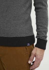 Suéter de punto a rayas negras y grises con dobladillo negro acanalado, que presenta una etiqueta en la esquina inferior izquierda. El usuario lleva pantalones beige y tiene una mano posicionada cerca.