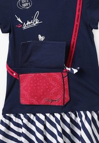 Laivastonsininen puuvillainen T-paita, jossa on punainen tasku ja raidallinen hame. Sisältää leikkisän tekstigrafiikan ja kontrastiset kuviot. Punaiset yksityiskohdat rusetilla.