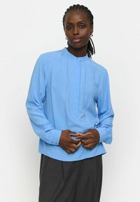 Bluză lungă cu mâneci lungi, de culoare albastru deschis, cu guler înalt, față plisată și manșete cu nasturi, realizată dintr-un material neted cu textură subtilă.